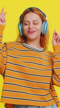 Bluz giymiş neşeli beyaz kadın kulaklıkla müzik dinliyor ve diskoda dans ediyor. Eğlenceli, jestli eller yapıyor. Sarı arka planda izole edilmiş genç bir kız.