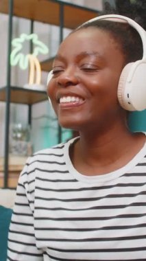 Afro-Amerikalı siyahi kadın dans ediyor, kablosuz kulaklıklarla müzik dinliyor rahat oturma odasında kanepede. Rahatlamak, gülümsemek, favori şarkıyı dinlerken eğlenmek. Ev hayatı, kız evde.