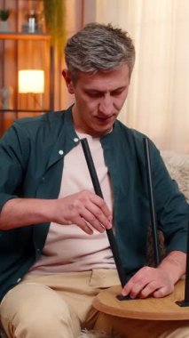 Evdeki genç adam sakin bir odak ile küçük ahşap masa onarıyor, ilerleme ve çabadan memnun görünüyor. Kanepede oturan beyaz adam işini yaparken ve mobilyaları tamir ederken konsantre olmuş görünüyor.