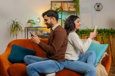 Evdeki kanepede arka arkaya akıllı telefonları kullanarak sırtüstü oturan neşeli ve çeşitli çiftlerin yan görüntüsü. Genç Hintli aile evdeki koltukta cep telefonu ararken konuşuyor.