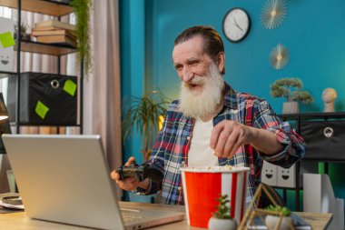 Bilgisayarda bilgisayar oyunu oynarken patlamış mısır yiyen heyecanlı Kafkasyalı yaşlı bir adam. Boş vakitlerini ofis masasında geçiriyor. Mutlu Bağımsız Son Sınıf Yetişkin Büyükbaba video oyunları oynuyordu..