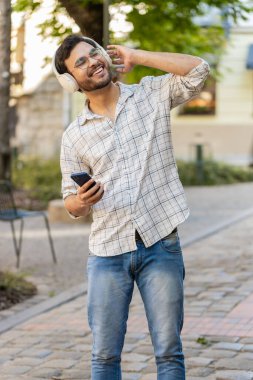 Kablosuz kulaklıklı mutlu genç turist dışarıda akıllı telefondan en sevilen enerjik disko müziğini dinliyor. Kentsel güneşli şehir parkında duran Hintli turist..