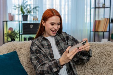 Evde heyecanlı bir şekilde oyun oynayan genç bir kadın. Ekrana vuruyor ve mutlu zaferi kutluyor. Kafkasyalı kızıl saçlı kız kanepede sevinçle gülümsüyor, başarının ve duygusal oyun anının tadını çıkarıyor.