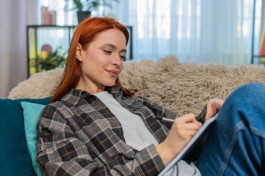 Evdeki genç kadın kanepeye oturup, düşünceli yüzüyle günlüğe yazarak gelecekle ilgili planlarını hayal ediyor. Kafkasyalı kızıl kız rahat, derin duygularla yansıtıyor, hedefleri ve niyetleri belgeliyor.