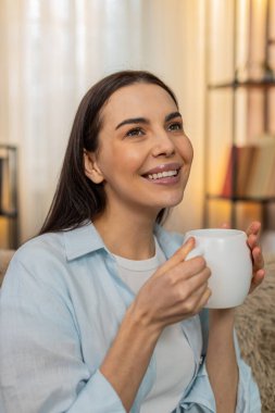 Genç ve mutlu bir kadın elinde çay fincanıyla mutlu bir şekilde aromasını soluyor, rahatlatıyor ve huzurlu bir sessizliğin tadını çıkarıyor. Kanepede tatmin olmuş, sakin duygular, rahatlık ve sükunet ifade eden beyaz bir kız.