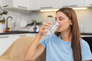 Evde su içen genç bir kadın susuzluğunu korur ve iyi bir amacı olan sağlıklı bir alışkanlığı izler. Kanepede oturan beyaz kız filtre sistemi kullanıyor. Gülümsemeler, içtikten sonra ferahlıyor ve sakinleşiyor..
