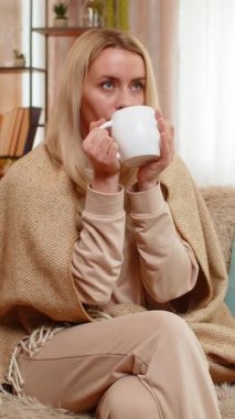 Genç, yetişkin bir kadın sıcak bir battaniyeye sarılmış, sıcak bir çay tutuyor ve sakin bir şekilde diziyi seyrediyor. Sarışın kız düşünceli görünüyor, sıcak içki ve konforla huzurlu bir anın tadını çıkarıyor.