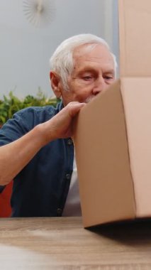 Evde kulaklıklarla teslimat paketini açan mutlu kıdemli adam. Gülümseyen büyükbaba alışverişçisi, çevrimiçi dükkan müşterisi karton kutu açıyor hızlı posta yoluyla satın alma hediyesi alıyor.