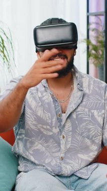 Hintli adam sanal gerçeklik teknolojisi VR uygulama kulaklığı kullanarak 3D video oyunu simülasyonu yapıyor ve modern ev dairesinde film izliyor. Kanepede oturan gözlüklü adam..