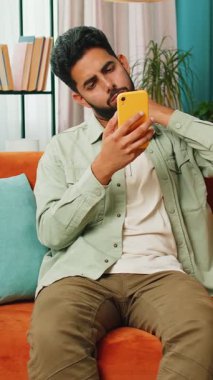 Hüzünlü Arap adam akıllı telefon daktilo kullanıyor, beklenmedik piyango sonuçlarını kaybediyor, kötü haber, servet kaybı, başarısızlık, teslim tarihi, virüs. Genç Hintli adam kanepede oturuyor.