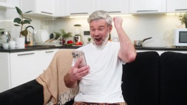 Evde orta yaşlı bir adam akıllı telefondan mutlu haberler okur ve online oyun kutlarken güler başarılı olur neşeli ruh hali kazanır. Kanepede oturan olgun bir adam gülümseyerek yumruk zaferini kaldırıyor..