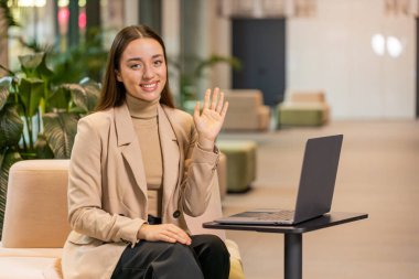 Ofis lobisinden uzaktan çekilmiş çevrimiçi konferans görüntülü konuşmasında gülümseyen serbest çalışan genç iş kadınının portresi. Koridorda dizüstü bilgisayarı olan resmi elbiseli bir kız..