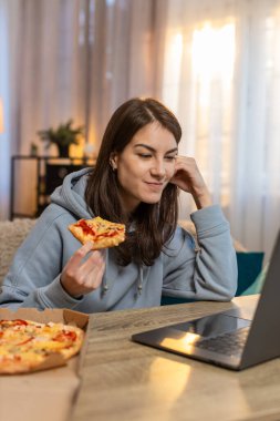 Genç, beyaz bir kadın lezzetli abur cubur pizzaları yerken masada oturmuş film izliyor ya da dizileri oturma odasında dizide izliyordu. Günlük kıyafetler içinde yetişkin bir kız. Hafta sonunun tadını çıkarıyor..