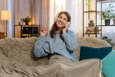 Kablosuz kulaklıkla müzik dinleyen neşeli genç bir kadın kanepede oturmuş ritme göre hareket ediyor. Evde müzik eşliğinde dans eden günlük kıyafetlerle neşeli bir kız. Dans müziğinin tadını çıkarıyorum. Yaşam Tarzları.