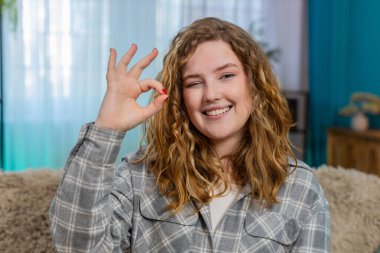 Evdeki genç kadın iyi el işareti gösteriyor. Kendine güvenen bir görünüm ve olumlu tavır sergiliyor. Kanepede gururla gülümseyen beyaz saçlı kız, duygusal güç ve kendinden emin duruş.