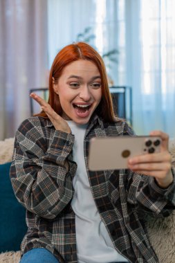 Evde heyecanlı bir şekilde oyun oynayan genç bir kadın. Ekrana vuruyor ve mutlu zaferi kutluyor. Kafkasyalı kızıl saçlı kız kanepede sevinçle gülümsüyor, başarının ve duygusal oyun anının tadını çıkarıyor.