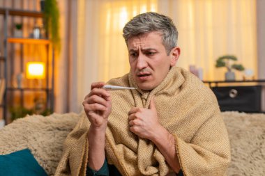 Evdeki genç adam endişeli görünürken vücut ısısını elektronik termometreyle ölçüyor. Kanepedeki beyaz adam endişeli ve rahatsız görünüyor. Hastalık ve belirsizlik belirtileri gösteriyor.