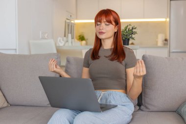 Evde genç bir kadın laptopçu işine ara veriyor ve derin nefes alarak meditasyon yapıyor. Kanepede oturan kızıl saçlı kız gözlerini kapatır ellerini kucağına koyar yavaşça nefes alır zihin rahatlar ve denge huzur bulur..