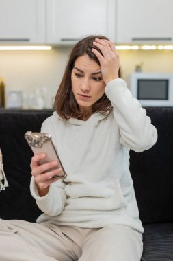 Evdeki genç kadın olumsuz telefon uyarısı okuyor. Neden böyle olduğunu fısıldıyor. Şok ve korku hissediyor. Kanepede oturan beyaz kız endişeli görünüyor. Akıllı telefonu sıkı tutuyor. Mesajı okuyor.