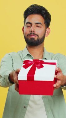 Pozitif gülümseyen Hintli adam doğum günü hediyelerini sunar ellerini uzatır, hediye paketi hediye ikramiyesi teklif eder, kutlama partisi düzenler, indirim indirimi yapar. Sarı arka planda izole edilmiş Arap adam.