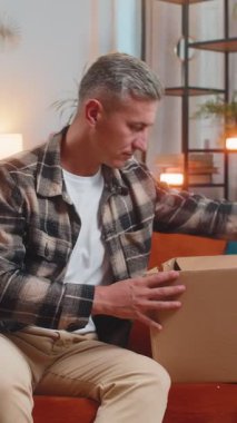 Evdeki kanepede oturan mutlu genç adam teslimat paketini açıyor. Gülümseyen memnun erkek alışverişci çevrimiçi dükkan müşterisi karton kutu açıyor hızlı posta yoluyla satın alma hediyesi alıyor.