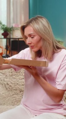 Evde orta yaşlı hamile bir kadın pizza kutusunu açar, aroma ısırıkları solur, akşam yemeğinin tadını çıkarır. Kanepede sırıtan beyaz bir kız. Hamilelik boyunca rahatça yemek yiyerek peynirin tadını çıkarıyor..