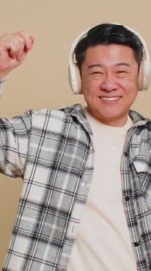 Orta yaşlı mutlu Asyalı adam kulaklıkla müzik dinliyor, dans ediyor, eğleniyor, el hareketleriyle kutlama yapıyor. Olgun yetişkin adam bej arka planda kapalı