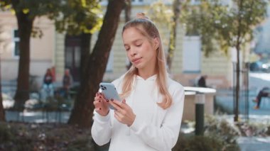 Mutlu, gülümseyen, beyaz bir kadın internet sosyal medya uygulamasında internette sohbet ederek akıllı telefon yazıları yazıyor. Genç kız turist şehir caddesinde duruyor..