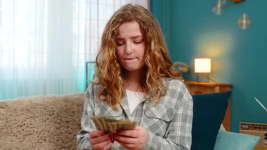 Evde elinde dolarlarla para sayan ve maddi durumu duygusal olarak düşünen genç bir kadın. Kanepedeki beyaz saçlı kız tatmin olmuş ya da endişeli görünüyor, kişisel bütçe yansıması.