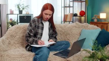 Evde genç bir kadın odaklanarak, başını sallayarak ve not defterine notlar yazarak çevrimiçi derse katılıyor. Kanepede oturan beyaz saçlı kız aktif olarak öğreniyor, eğitime ve sanal sınıfa odaklanıyor.