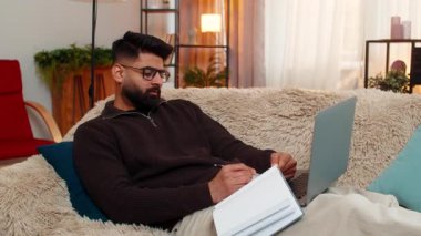 Evde oturan Hintli adam dizüstü bilgisayar okuyup öğrenmeye odaklanmış bir şekilde defterine dikkatlice yazıyor. Kanepedeki Arap adam sakince çalışıyor. İnternetteki sınıfa hazırlanıyor. Dikkat, çaba ve özveri ile.