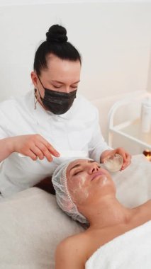 Güzellik uzmanı, yetişkin kadın yüzüne spatulayla nemlendirici tıbbi kimyasallar soyarak krem maske sürüyor. Dişi cilt bakımı prosedürü. Yüz kozmetolojisi cilt yenilenmesi. Dikey