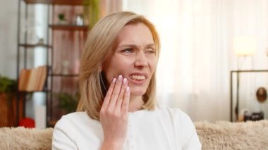 Orta yaşlı bir kadın, diş ağrısından ürkmüş bir vaziyette elinde yanağıyla kanepesinde oturuyor. Yetişkin hasta kadın gergin bir yüz ve vücut diline sahip keskin bir ağrı hissediyor. Rahatlamak ve sakinleşmek istiyor..