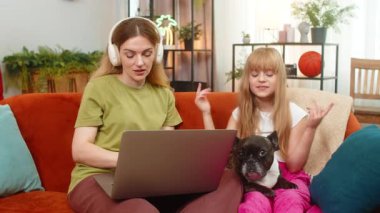 Evde Fransız bulldog 'u olan anne-kız kablosuz kulaklık kullanıyor dizüstü bilgisayar çalışırken müzik dinliyor. Kanepede köpekli bir kadın ve kız. Step ritmi notaları eşliğinde şarkı söylüyorlar. Odaklan.