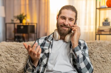 Genç adam evde kablosuz kulaklıklarla oturuyor, telefonları kaydırıyor ve müzik dinliyor. Kanepedeki adam gelen arama için kulaklıkları çıkarıyor, neşeli, neşeli bir sohbet başlatıyor.