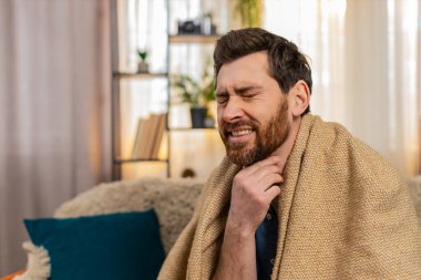 Evdeki genç adam kanepeye oturur, battaniyeye sarınır, öksürür ve acı içinde boğazını tutar. Kafkasyalı adam kaşlarını çatmış, yutkunmaya çalışmış, muhtemelen boğaz ağrısı ya da grip belirtileri göstermiş.