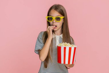 Genç kız mutlu bir şekilde patlamış mısır yiyor, televizyonda eğlenceli bir çizgi film izliyor, neşeli bir şekilde gülüyor, geniş bir kesime gülümsüyor, eğlenceli animasyon sahnelerinden haz alıyor. Pembe arka plandaki okul çocuğu.