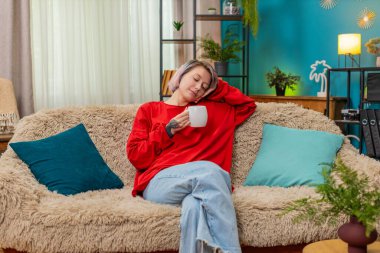 Evde sakince kahve içen genç bir kadın, rahatlamış, tatmin olmuş, huzurlu bir zevk ve rahatlık ifade ediyor. Mutlu kız sessizce gülümsüyor, rahatlıyor, kanepede içecekle keyifli vakit geçiriyor..