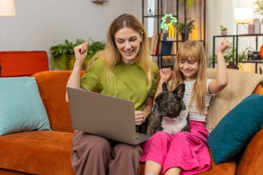 Evde Fransız bulldog 'u olan anne ve kızı dizüstü bilgisayarlarını bitirip zafer testi sonucu ponpon alkışı aldılar. Kanepede köpekli bir kadın ve kız zaferi kutluyor. İyi haber gülüşü ödülü.