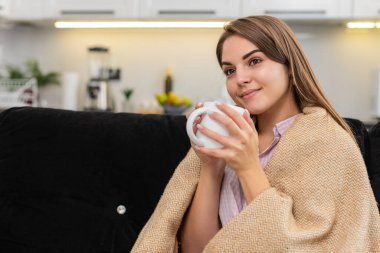 Evdeki genç kadın bitkisel çay içiyor ve kupanın üzerinde ellerini ısıtıyor ve yavaş yavaş nefes alıyor ve sakinleşiyor. Kanepede oturan beyaz kız gülümsüyor ve sessizliğin tadını çıkarıyor ve dinleniyor ve sıcak bir ruh hali içinde dinleniyor ve rahatlıyor.