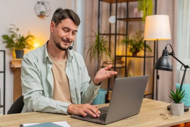 Kulaklık takan genç bir adam, serbest çalışan, çağrı merkezi ya da destek operatörü yardım hattı, müşteri ya da iş arkadaşı iletişim desteği ile konuşuyor. Ofis masasında dizüstü bilgisayar kullanan bir adam. Yaşam biçimi.