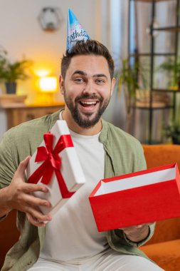 Mutlu genç adam doğum günü şapkası takıyor ve elinde kurdeleyle kutluyor. Beyaz tenli esmer erkek, parti açılışını kutluyor. Kanepede, toplantı odasında hediyeler sunuyor..