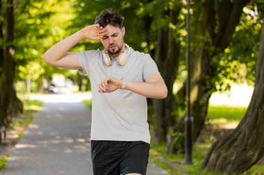 Yoğun kardiyo eğitimi. Genç, aktif bir erkek koşucu parkta koşuyor ve akıllı adımlara bakıyor, yoruluyor ve yavaşlıyor. Sporcu, dışarıda ağaçlarla kalp atışlarını kontrol ediyor..