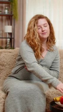 Hamile, genç bir kadın, bir eliyle karnına hafifçe dokunarak kanepeye oturur ve taze elmadan bir ısırık alır ve besleyici atıştırmalıkların tadını çıkarır. Kız huzur ve tatmin hissediyor sağlıklı seçimi kucaklıyor.