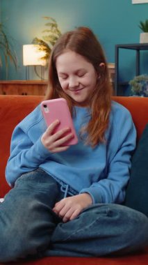 Neşeli genç kız, internet üzerinden akıllı telefon sosyal medya uygulamalarında rahatlama filmi izlerken mesajlarını paylaşıyor. Oturma odasında cep telefonu kullanan beyaz bir kız çocuk kanepede oturuyor..