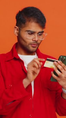 Kredi kartı ve akıllı telefon kullanan mutlu genç Hintli adam harcamaları transfer ederken, online alışveriş yaparken, yemek siparişi verirken. Arap adam turuncu stüdyo arka planında izole edilmiş.