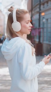 Kablosuz kulaklıkla gezen mutlu genç kadın turistin arka görüntüsü, açık havada akıllı telefondan en sevilen enerjik disko müziğini dinliyor. Güneşli şehir sokağının yanından geçen beyaz bir genç..