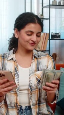 Hintli genç bir kadın bir elinde akıllı telefonuyla kanepede oturuyor, diğerinde de nakit paraları sayıyor. Arabistanlı kız kişisel harcamalar planlıyor, dikkatli bütçe ayırıyor ya da online aktarımlar yapıyor