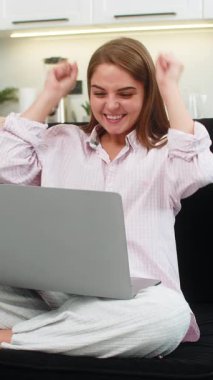 Evdeki genç kadın dizüstü bilgisayar güncellemelerini okuyor ve kazanma haberleri üzerine sevinçle kollarını kaldırıyor. Kanepede oturan beyaz kız zaferi kutluyor neşeli bir ruh hali ve iyi sonuçlara canlı bir tepki gösteriyor.