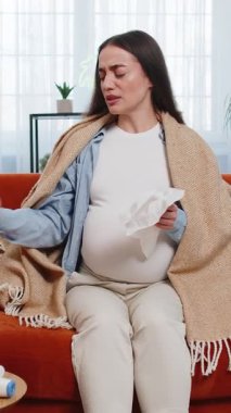 Oturma odasındaki kanepede otururken, battaniyeye sarılmış, mendile burunlarını sümküren hamile, sağlıksız beyaz kadın. Müstakbel annem evde burun tıkanıklığı ve soğuk algınlığı semptomlarından bıktı..